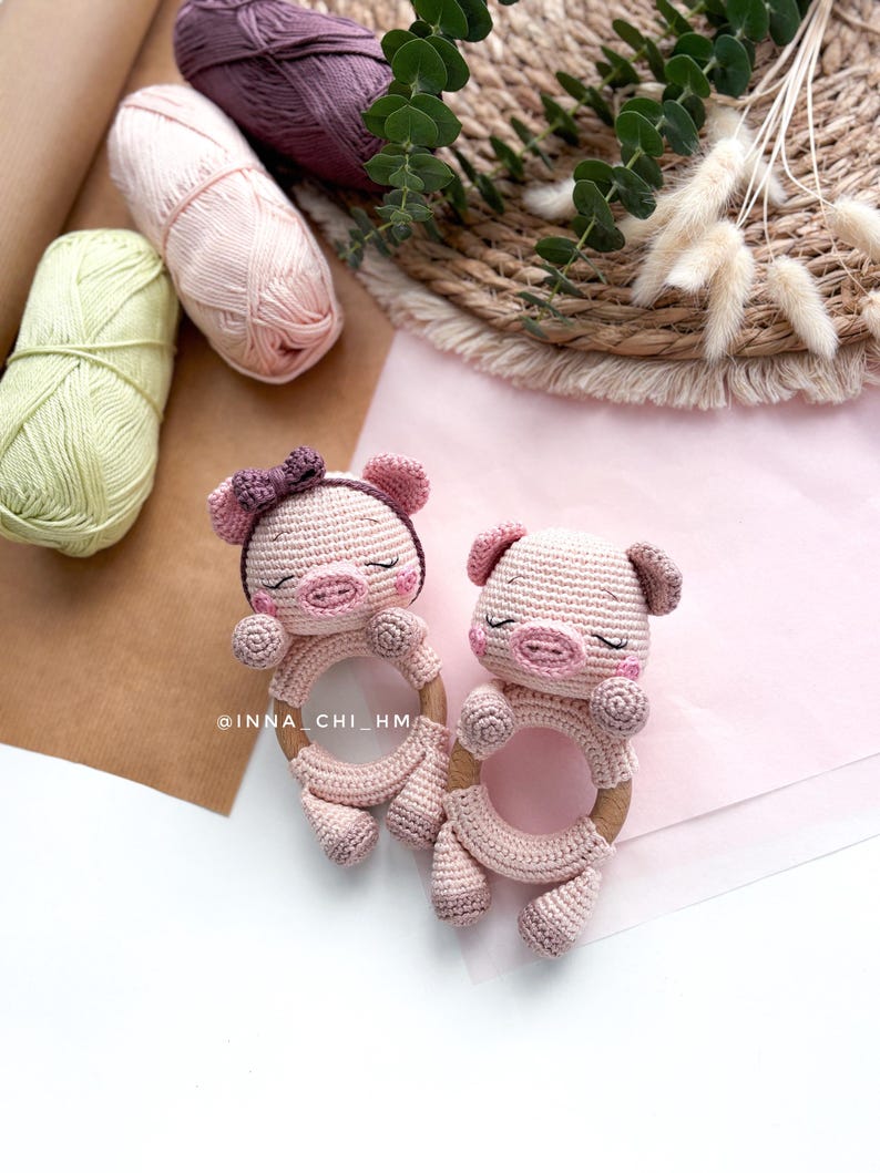 May include: Two handmade crochet piglet baby toys in light pink with wooden rings. The pigs have closed eyes and small ears. The background includes skeins of yarn in green, pink, and purple, along with decorative elements.