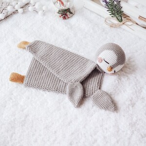 Peut inclure: Un jouet en crochet de pingouin gris avec un visage blanc et des joues roses. Le pingouin est couch&eacute; sur le c&ocirc;t&eacute;, les ailes d&eacute;ploy&eacute;es. Le jouet est sur une surface blanche et moelleuse.