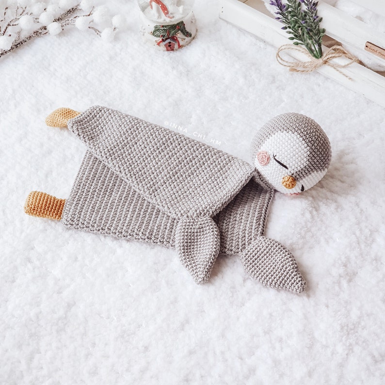 Peut inclure: Un jouet en crochet de pingouin gris avec un visage blanc et des joues roses. Le pingouin est couch&eacute; sur le c&ocirc;t&eacute;, les ailes d&eacute;ploy&eacute;es. Le jouet est sur une surface blanche et moelleuse.