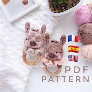 May include: Two handmade crochet bunny rattles in shades of brown and pink, each with a wooden ring. The bunnies have closed eyes and a ruffled collar. The image includes the text "PDF PATTERN".