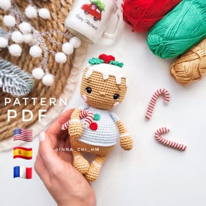 Op de afbeelding: Haakpatroon voor een amigurumi-peperkoekman speelgoed in PDF-formaat. Het patroon bevat instructies voor het maken van een peperkoekman met een wit shirt, een rode snoepstok en groene hulstbladeren. Het patroon is beschikbaar in het Engels, Spaans en Frans.