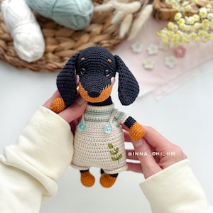 May include: A handmade crochet dachshund dog wearing a cream-colored dress with blue buttons and a green leaf design. The dog has black fur, orange paws, and a tan snout. The background includes yarn and flowers.