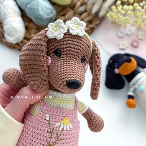 May include: A crocheted brown dachshund dog wearing a pink overall with a daisy design. The dog has a white flower crown and a yellow and white striped shirt. A second crocheted dog is visible in the background.