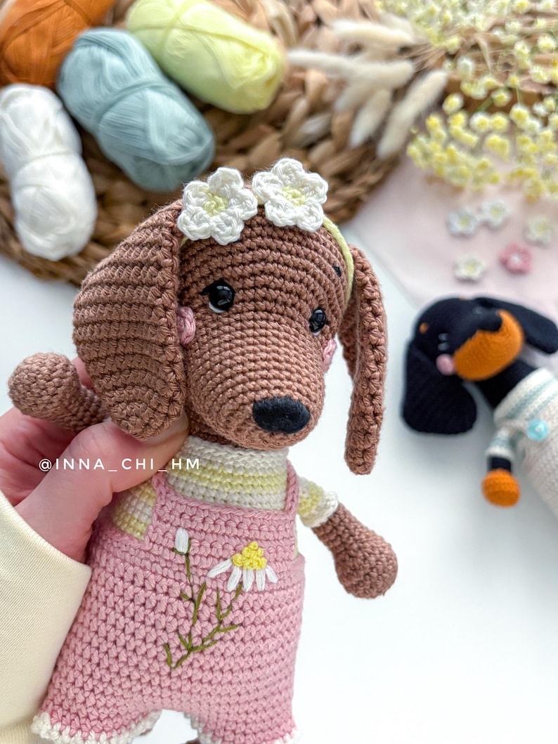 May include: A crocheted brown dachshund dog wearing a pink overall with a daisy design. The dog has a white flower crown and a yellow and white striped shirt. A second crocheted dog is visible in the background.