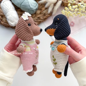 May include: Two crocheted dog figurines. One is brown with a white flower and pink overalls. The other is a black and tan dachshund with a white shirt and orange accents. The figurines are handmade and detailed.