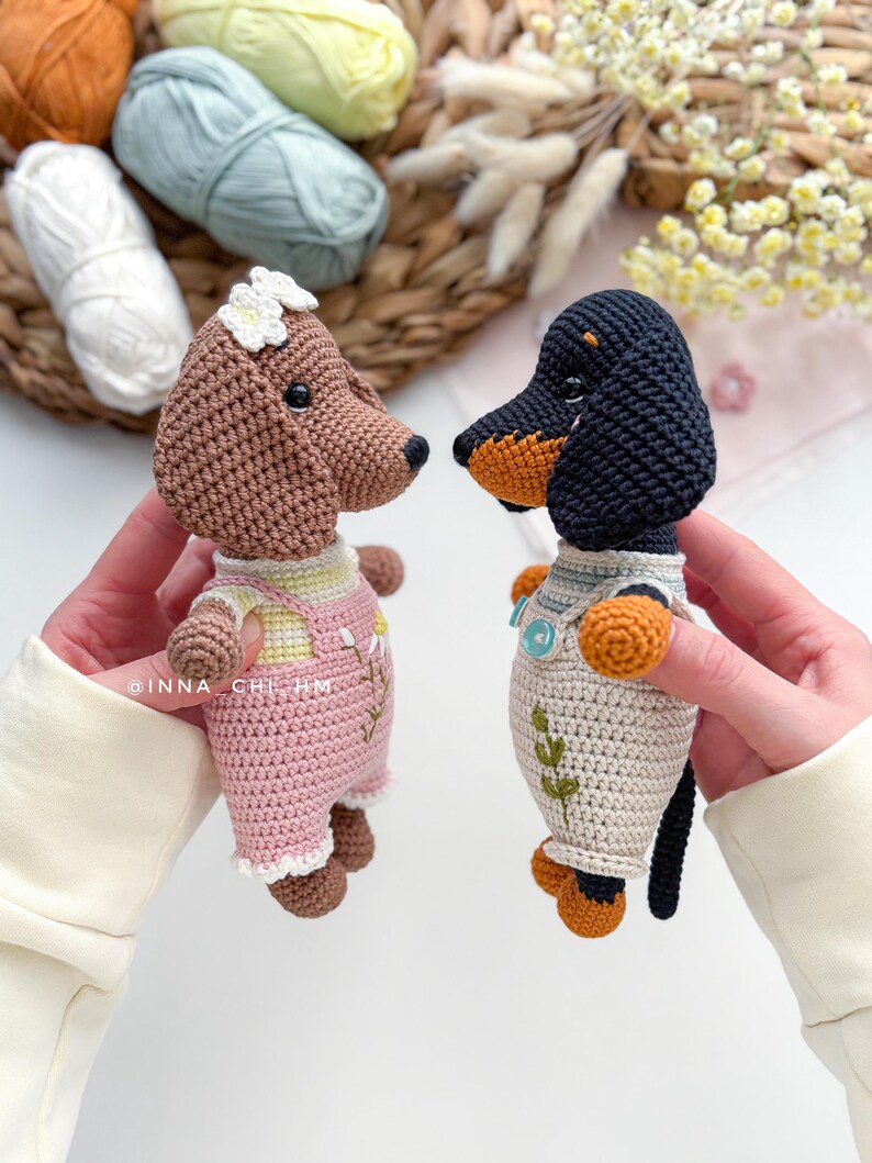 May include: Two crocheted dog figurines. One is brown with a white flower and pink overalls. The other is a black and tan dachshund with a white shirt and orange accents. The figurines are handmade and detailed.