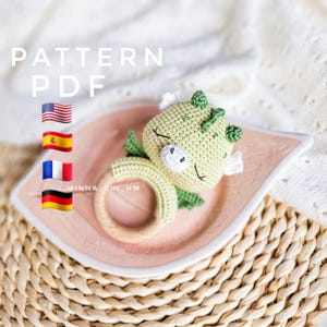 以下が含まれることがあります： 木製の輪が付いた、かぎ針編みのドラゴンのおしゃぶり。ドラゴンは緑色で、薄い緑色の腹をしています。目は閉じられていて、かわいい顔をしています。おしゃぶりは、葉っぱのデザインが施されたピンク色の皿の上に置かれています。