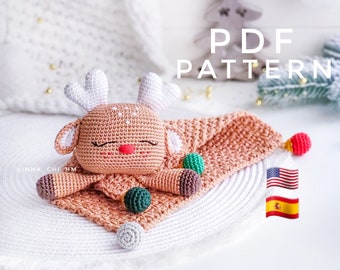 Puede incluir: Patrón de crochet PDF para un reno marrón con la nariz roja y los ojos cerrados, acostado sobre una manta marrón y blanca con bolas verdes y marrones. El patrón está disponible en inglés y español.
