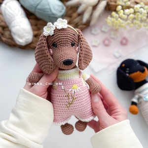 May include: A handmade crochet dachshund dog wearing a pink overall with a daisy design. The dog has brown fur, floppy ears, and a white flower crown. The text "@INNA_CHI_HM" is visible on the dog's arm. Another crochet dog is in the background.