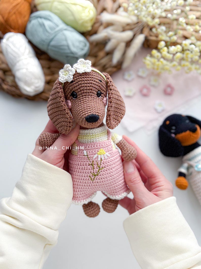 May include: A handmade crochet dachshund dog wearing a pink overall with a daisy design. The dog has brown fur, floppy ears, and a white flower crown. The text "@INNA_CHI_HM" is visible on the dog's arm. Another crochet dog is in the background.