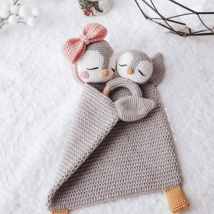 Peut inclure: Une couverture b&eacute;b&eacute; en crochet gris avec deux jouets en forme de pingouin en crochet. Les jouets pingouins tiennent un anneau en bois. La couverture a un d&eacute;tail en crochet jaune en bas.