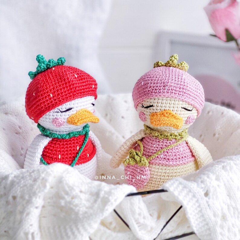 Peut inclure: Deux poup&eacute;es canards crochet&eacute;es &agrave; la main dans un panier blanc. L'un porte un chapeau et un gilet fraise rouges. L'autre a un chapeau et un gilet roses, avec un petit sac &agrave; main fraise. Les deux ont les yeux ferm&eacute;s et un bec orange.