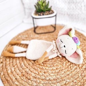 May include: A crocheted white bunny with pink ears and a flower crown is lying on a woven straw mat. The bunny has closed eyes and a sweet expression.