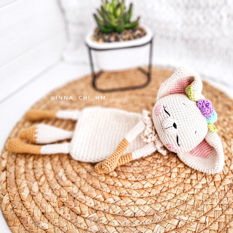 May include: A crocheted white bunny with pink ears and a flower crown is lying on a woven straw mat. The bunny has closed eyes and a sweet expression.