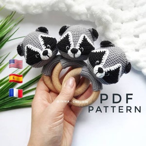 May include: Three crocheted raccoon toys attached to a wooden teething ring. The ring is held in a hand. The text "PDF PATTERN" is visible in the image.