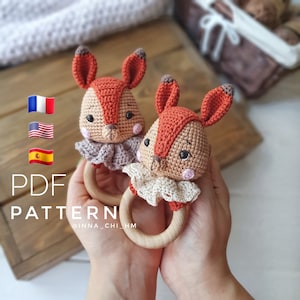 Puede incluir: Dos juguetes de sonajero de zorro de crochet con anillos de madera. Los zorros son marrones y naranjas con mejillas rosadas y ojos marrones. Los juguetes están sostenidos en las manos de una persona.