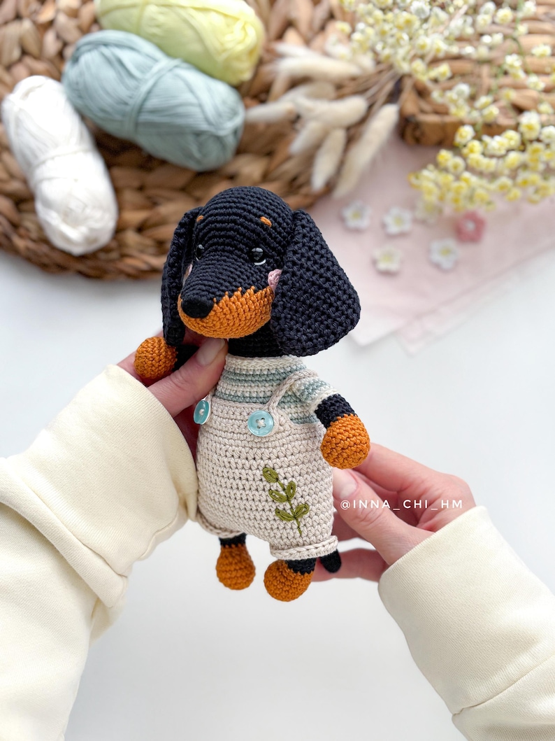 May include: A handmade crochet dachshund dog with a black body, tan face and paws, and wearing cream-colored overalls with a green leaf design. The dog is held by hands, with yarn and flowers in the background.