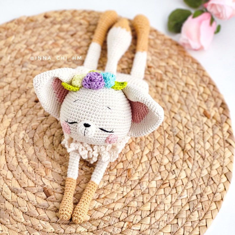 May include: A crocheted white bunny with long legs and a flower crown. The bunny is lying on a woven straw mat.