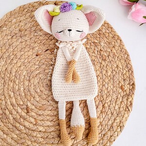 May include: A crocheted stuffed animal in the shape of a fox with a flower crown. The fox is white with light brown accents and is lying on a woven straw mat.