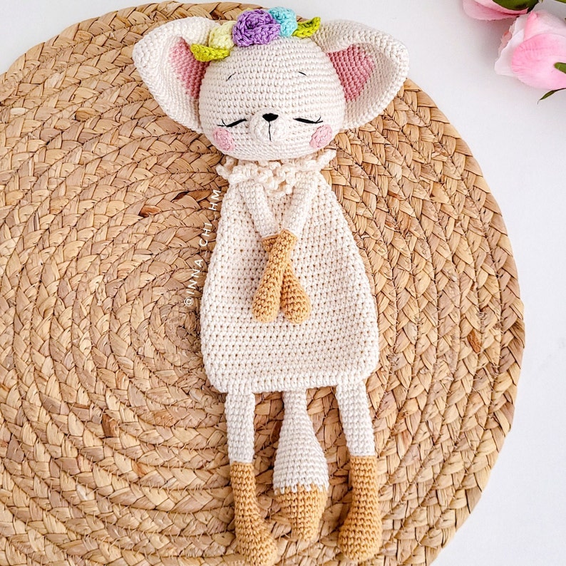 May include: A crocheted stuffed animal in the shape of a fox with a flower crown. The fox is white with light brown accents and is lying on a woven straw mat.