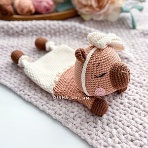 May include: A crocheted brown and white pig with a white bow, lying on a pink knitted blanket. The pig has a white stripe around its face and pink cheeks.