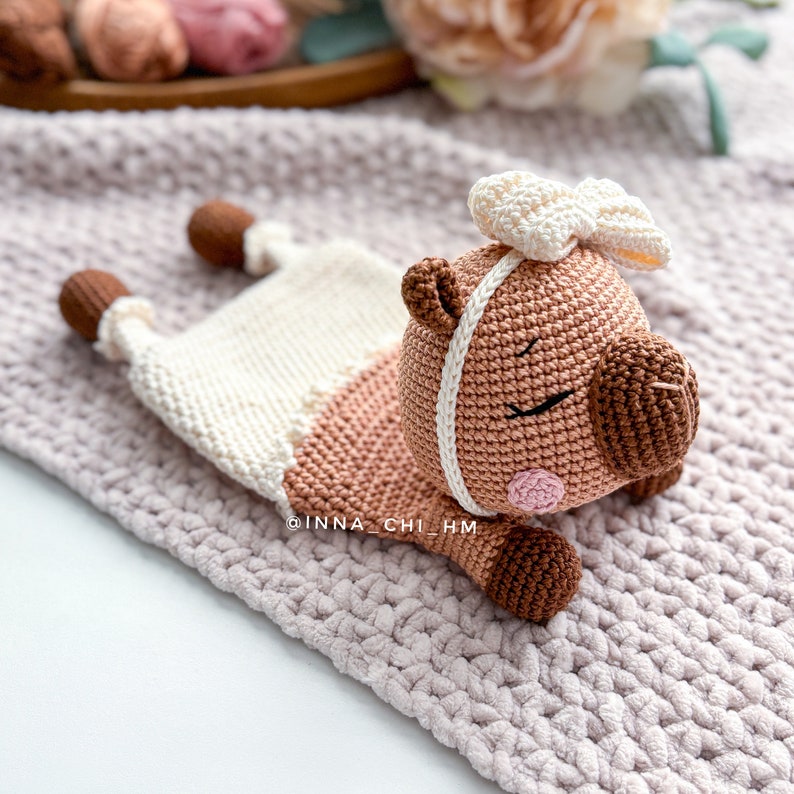 May include: A crocheted brown and white pig with a white bow, lying on a pink knitted blanket. The pig has a white stripe around its face and pink cheeks.