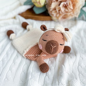 May include: A crocheted brown and white llama toy with a white bow. The llama is lying on a white crocheted blanket.