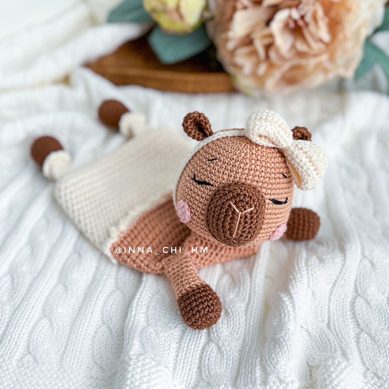 May include: A crocheted brown and white llama toy with a white bow. The llama is lying on a white crocheted blanket.