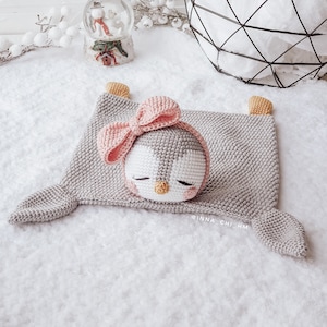 Peut inclure: Une couverture b&eacute;b&eacute; en crochet gris avec un jouet en crochet de pingouin avec un n&oelig;ud rose et les yeux ferm&eacute;s. La couverture a deux longs bras et une petite t&ecirc;te.