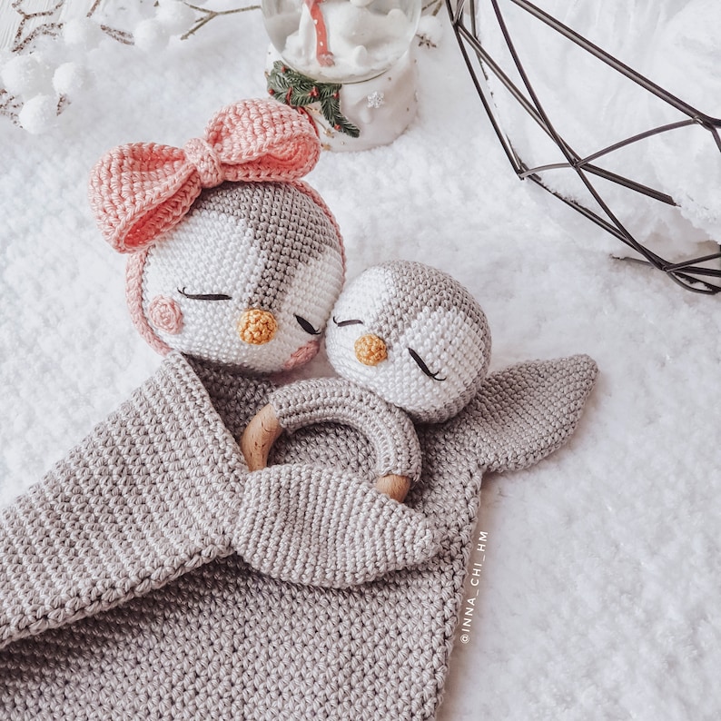 Peut inclure: Une couverture b&eacute;b&eacute; en crochet gris avec un n&oelig;ud rose et deux jouets en forme de pingouin en crochet. Le plus grand pingouin a un anneau en bois attach&eacute; &agrave; son aile. Le plus petit pingouin est nich&eacute; dans la couverture.