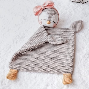Peut inclure: Une couverture de b&eacute;b&eacute; en forme de pingouin en crochet gris avec un n&oelig;ud rose sur la t&ecirc;te et des pieds jaunes. La couverture est pos&eacute;e sur une surface blanche.