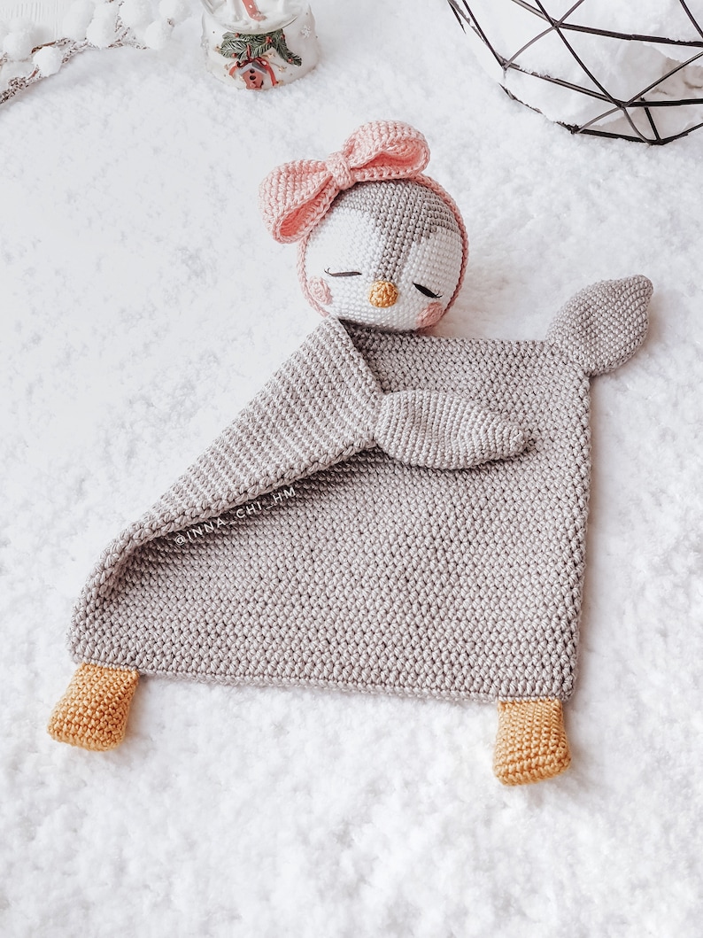 Peut inclure: Une couverture de b&eacute;b&eacute; en forme de pingouin en crochet gris avec un n&oelig;ud rose sur la t&ecirc;te et des pieds jaunes. La couverture est pos&eacute;e sur une surface blanche.