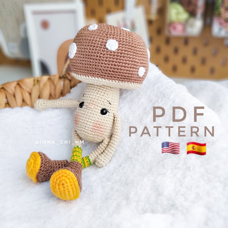 Op de afbeelding: Gehaakt amigurumi speelgoed in de vorm van een paddenstoel met een bruine dop, witte stippen en een beige lichaam. Het speelgoed heeft gele en bruine laarzen en groene strepen op de benen. De afbeelding bevat de tekst "PDF PATTERN" en vlaggen voor de Verenigde Staten en Spanje.