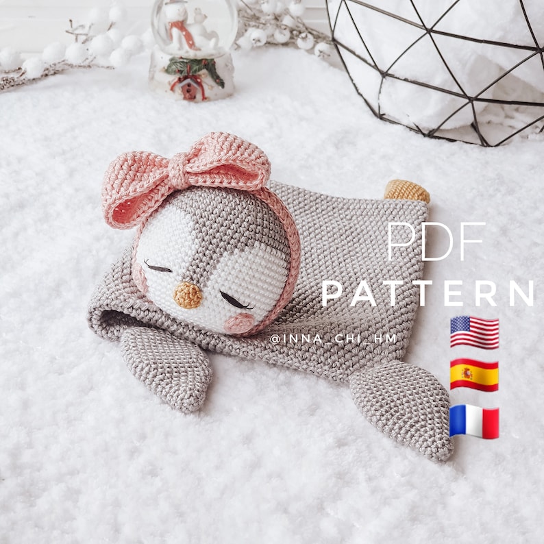Peut inclure: Un jouet en crochet amigurumi de pingouin avec un n&oelig;ud rose, couch&eacute; sur une surface blanche et moelleuse. Le jouet porte un bonnet en crochet gris. Le texte "PDF PATTERN" est visible sur l'image. L'image comprend &eacute;galement les drapeaux des &Eacute;tats-Unis, de l'Espagne et de la France.