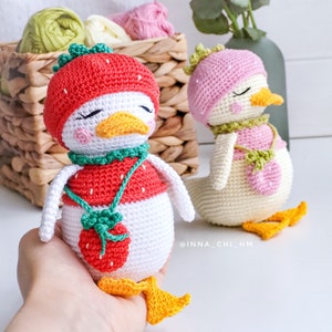 Peut inclure: Deux poup&eacute;es canard crochet&eacute;es, l'une tenue dans une main. Les canards sont blancs avec des becs et des pieds jaunes. L'un porte un chapeau et une chemise fraise rouges, l'autre un chapeau et une chemise roses. Les deux ont des accents verts et les yeux ferm&eacute;s.
