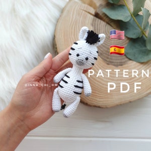 Puede incluir: Un juguete de peluche de cebra tejido a crochet con rayas negras y una crin negra. El juguete está siendo sostenido en una mano. El fondo incluye una superficie de madera, una bandera de Estados Unidos y una bandera de España. También se puede ver el texto "PATTERN PDF".
