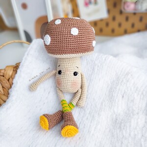 Op de afbeelding: Een gehaakt speelgoedpaddenstoel met een bruine dop, witte stippen en een beige lichaam. De paddenstoel heeft een schattig gezicht met zwarte ogen en een roze neus. Hij draagt ​​bruine laarzen met gele accenten en groen-gele gestreepte sokken. De paddenstoel zit op een wit deken.