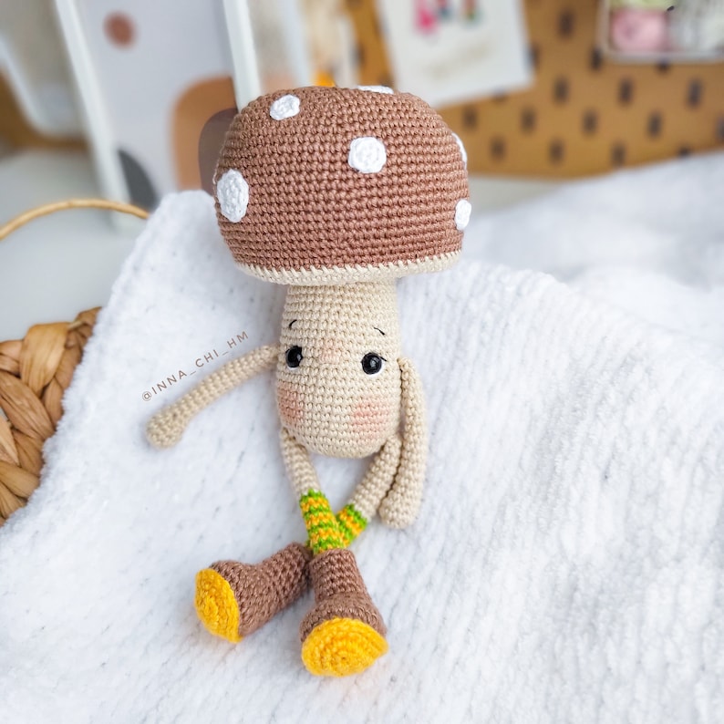 Op de afbeelding: Een gehaakt speelgoedpaddenstoel met een bruine dop, witte stippen en een beige lichaam. De paddenstoel heeft een schattig gezicht met zwarte ogen en een roze neus. Hij draagt ​​bruine laarzen met gele accenten en groen-gele gestreepte sokken. De paddenstoel zit op een wit deken.