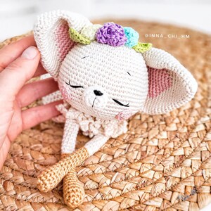 May include: A white crocheted bunny with pink ears, a flower crown with purple, blue, and green flowers, and brown legs. The bunny is lying on a woven straw mat. The text '@inna_chi_hm' is visible in the upper right corner of the image.