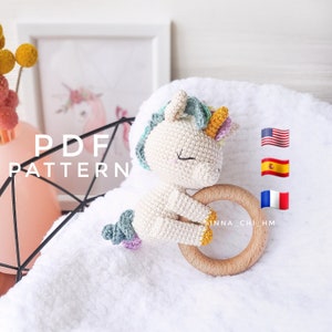 May include: Crochet unicorn amigurumi toy with a wooden teething ring. The unicorn is white with a rainbow mane and tail. The toy is on a white towel with the text "PDF PATTERN" and flags for the United States, Spain, and France.