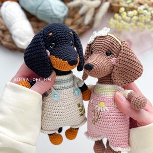 May include: Two handmade crochet dachshund dogs. One is black and tan, wearing a beige overall dress. The other is brown, wearing a pink dress with a daisy. Both have button eyes and are held up by a person.