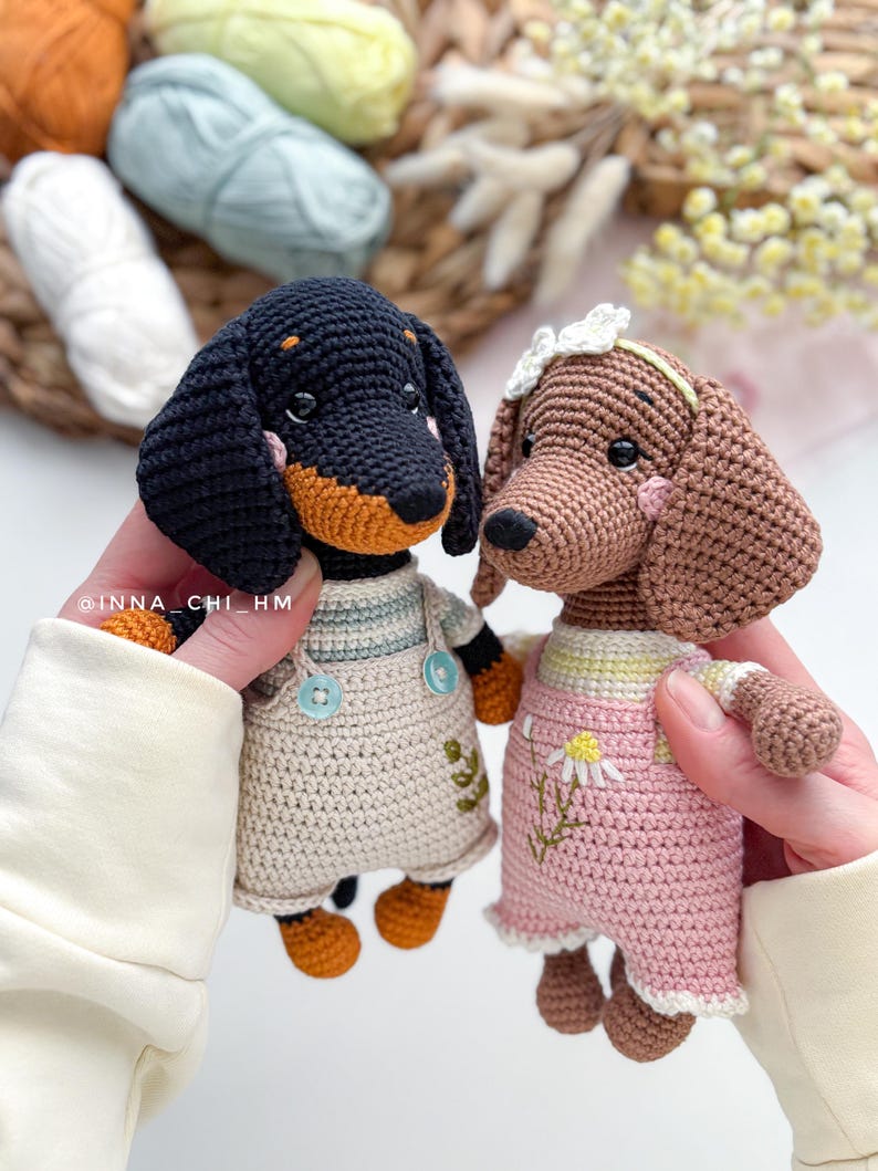 May include: Two handmade crochet dachshund dogs. One is black and tan, wearing a beige overall dress. The other is brown, wearing a pink dress with a daisy. Both have button eyes and are held up by a person.