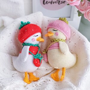 Peut inclure: Deux poup&eacute;es canard faites au crochet. L'une porte un chapeau fraise rouge et une tenue assortie, l'autre un chapeau et une &eacute;charpe roses. Les deux ont des becs et des pieds jaunes, sur fond blanc.