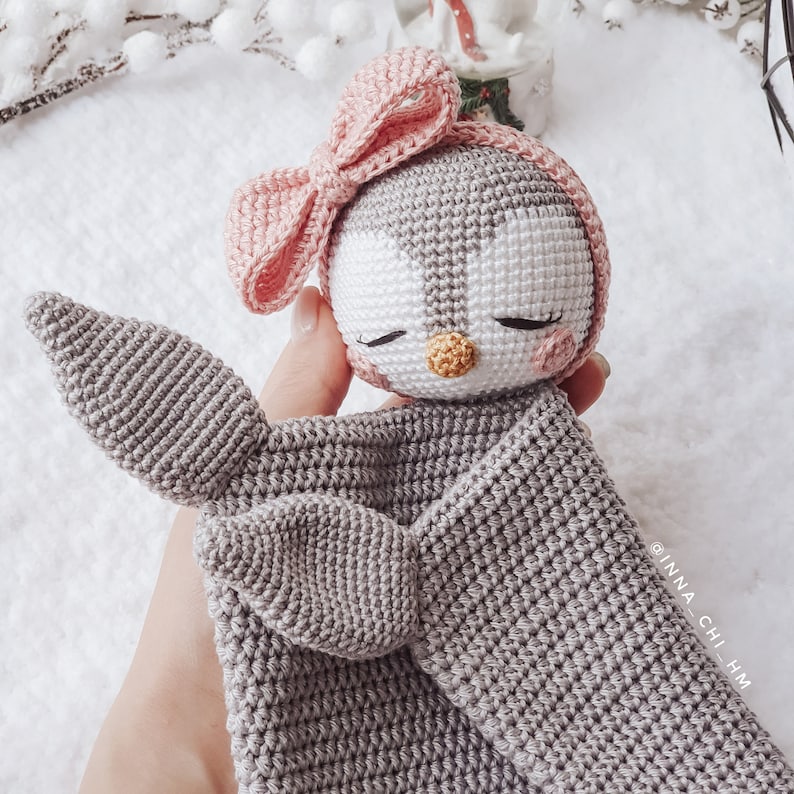 Peut inclure: Un pingouin en crochet gris avec un n&oelig;ud rose, tenant une couverture en crochet gris. Le pingouin a un visage blanc avec des yeux noirs et un bec jaune. La couverture a deux bras en crochet gris.
