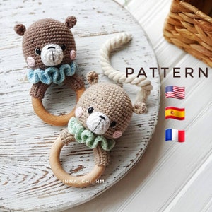 Puede incluir: Dos sonajeros de oso de crochet marrón con anillos de madera. Los osos tienen caras de color marrón claro con narices blancas y mejillas rosadas. Un oso tiene un volante azul y el otro un volante verde. La palabra "PATTERN" está escrita en letras negras sobre un fondo blanco. Las banderas de los Estados Unidos, España y Francia se muestran debajo de la palabra "PATTERN".