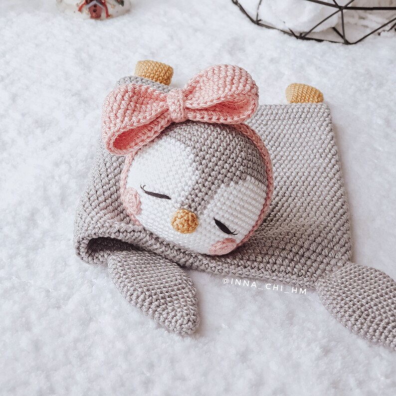 Peut inclure: Un jouet en crochet en forme de pingouin avec un n&oelig;ud rose, couch&eacute; sur une couverture en crochet grise. Le pingouin a un visage blanc avec des yeux noirs et un bec jaune.