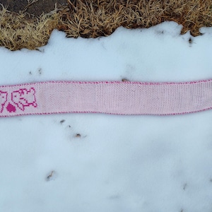 May include: A pink knitted headband with a heart and sheep design. The headband is light pink with a darker pink trim and design. The headband is lying on a bed of snow.