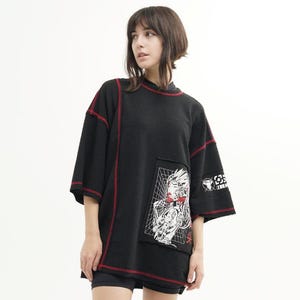 May include: Black oversized t-shirt with red stitching. It has a graphic patch with a detailed illustration. The sleeves are short and wide. The model is wearing black boots.
