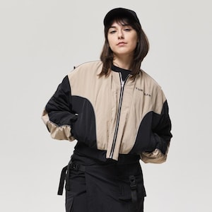 Könnte beinhalten: Eine Person trägt eine schwarze Baseballkappe, eine beige und schwarze Bomberjacke, eine schwarze Cargohose und schwarze Kampfstiefel. Auf der Jacke steht das Wort "TUR". Das Outfit ist ein moderner, urbaner Stil.