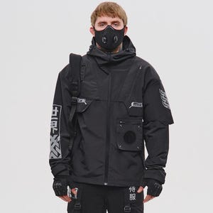 Può includere: Giacca tattica nera con più tasche, cappuccio e maschera. L'outfit include pantaloni cargo neri abbinati con cinghie e scarpe da ginnastica nere. Caratteri giapponesi bianchi sono stampati sulla giacca e sui pantaloni.